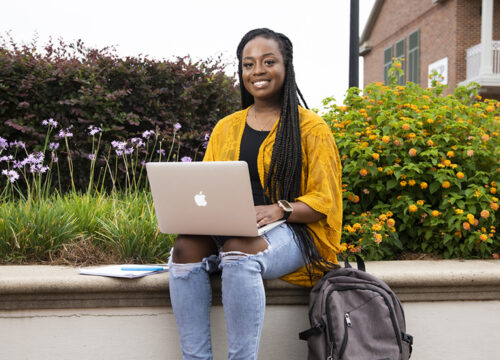 A student using a laptop while sitting on a concrete bench outdoors in front of bushes.