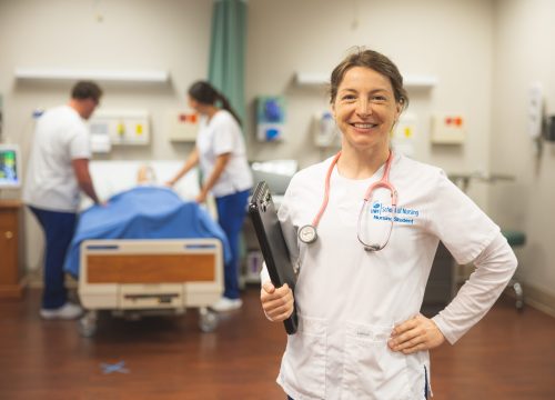 Nursing student with a stethoscope around her neck holds a clipboard in her right hand while two more nursing students work in the background