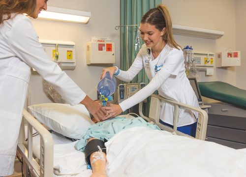 Two nursing students practicing respiratory therapy work in a hospital simulation lab setting.