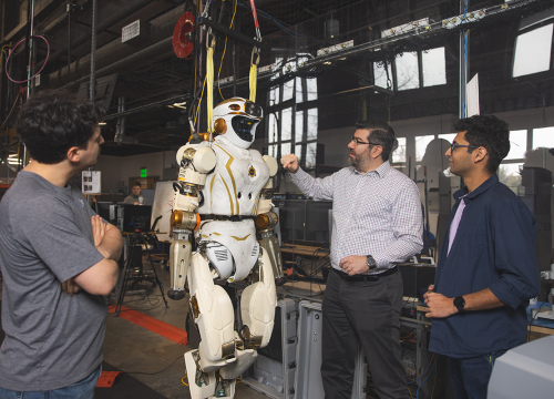 Three people look at a life-size robot in a lab